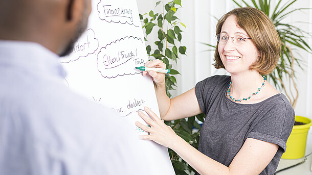 Eine Studienberaterin schreibt auf einem Flipchart und ein Student sieht zu