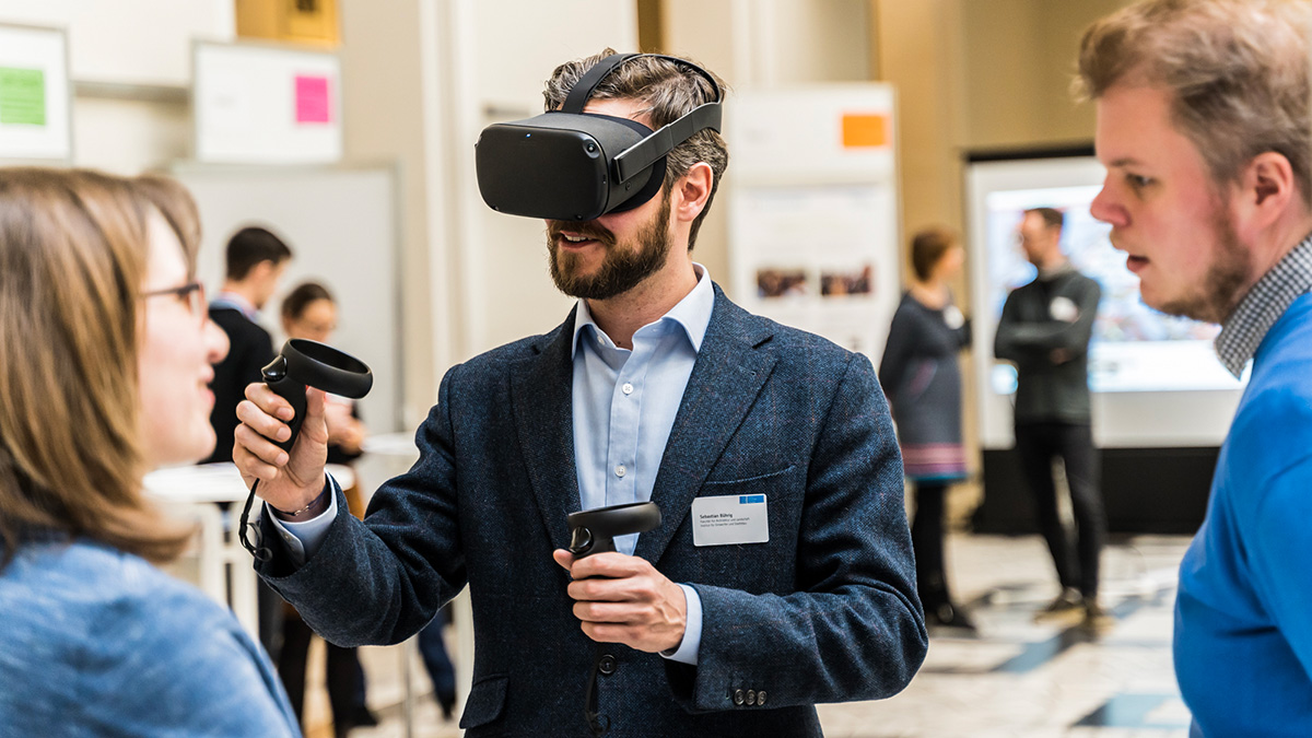Eine Person testet eine Brille zu virtueller Realität, daneben zwei weitere Personen im Austausch.