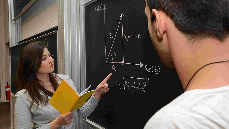 Eine Frau erklärt einem Mann eine mathematische Rechnung an der Tafel.