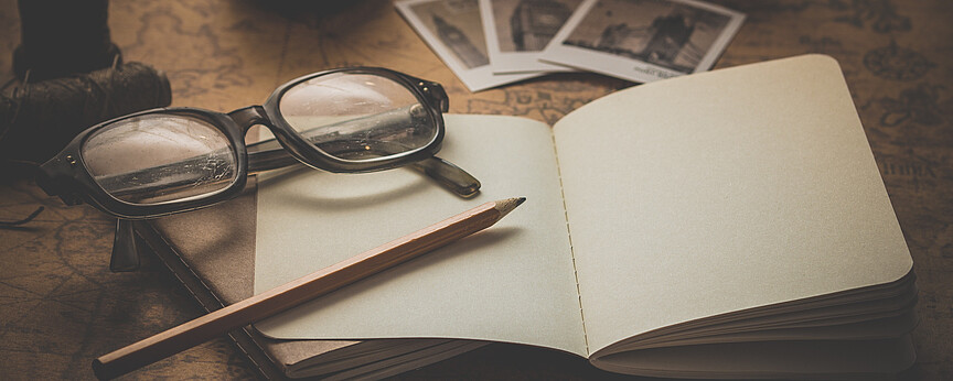 Auf einer antiken Weltkarte liegen ein aufgeklapptes Notizbuch, eine Brille und ein Bleistift. Im Hintergrund steht eine alte Kamera und drei Polaroid-Fotos.