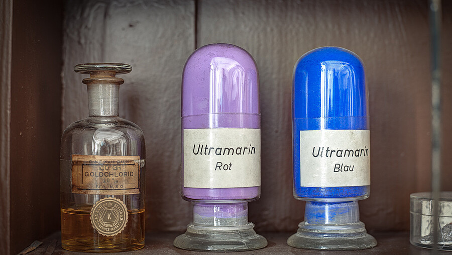 In einem Regal stehen Gefäße mit den Aufschriften "Goldohlorid", "Ultramarin Rot" und "Ultramarin Blau".