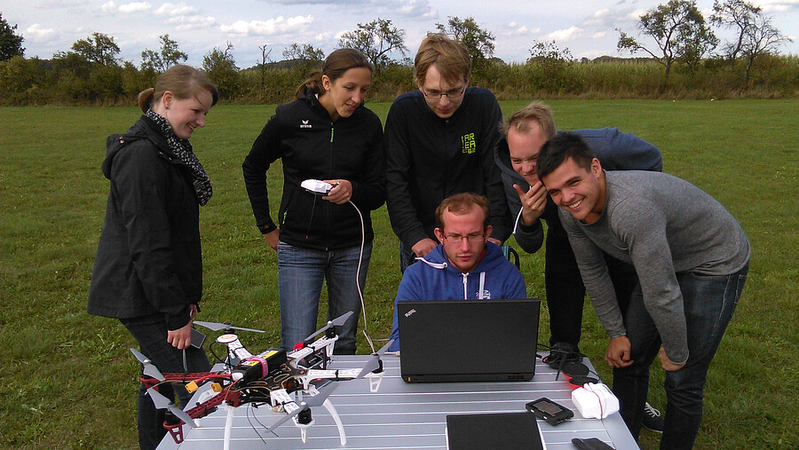 Six people work outside on a laptop and a drone.