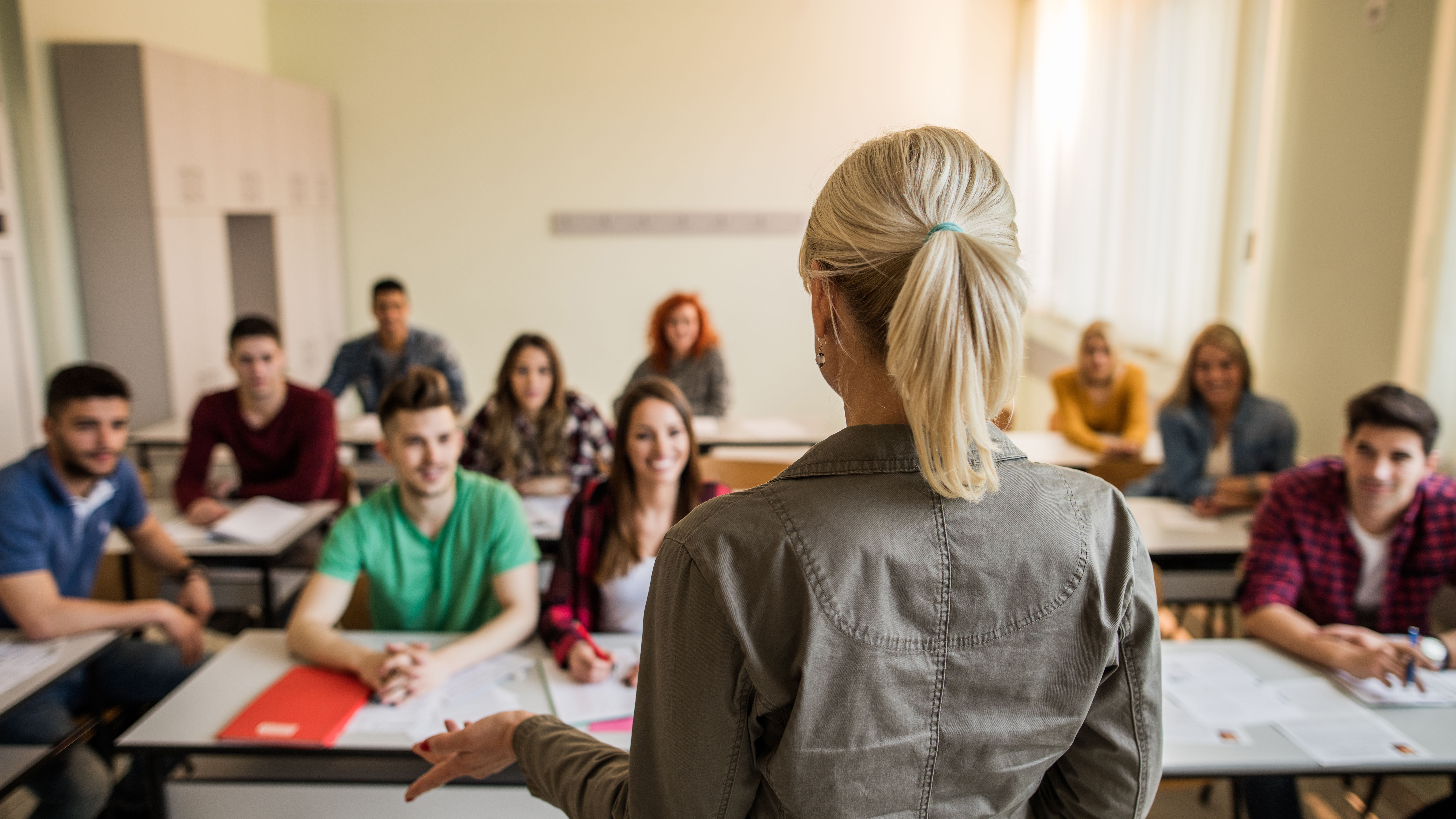 Im Vordergrund ist eine Lehrerin von der Hinterseite zu sehen und im Hintergrund eine Klasse, die ihr zuhört.
